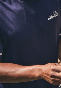 Námořnická modrá sportovní polokošile s krátkým rukávem, se bílými okraji a embosovaným logem na levé hrudi. Hladká textura, přiléhavý střih.