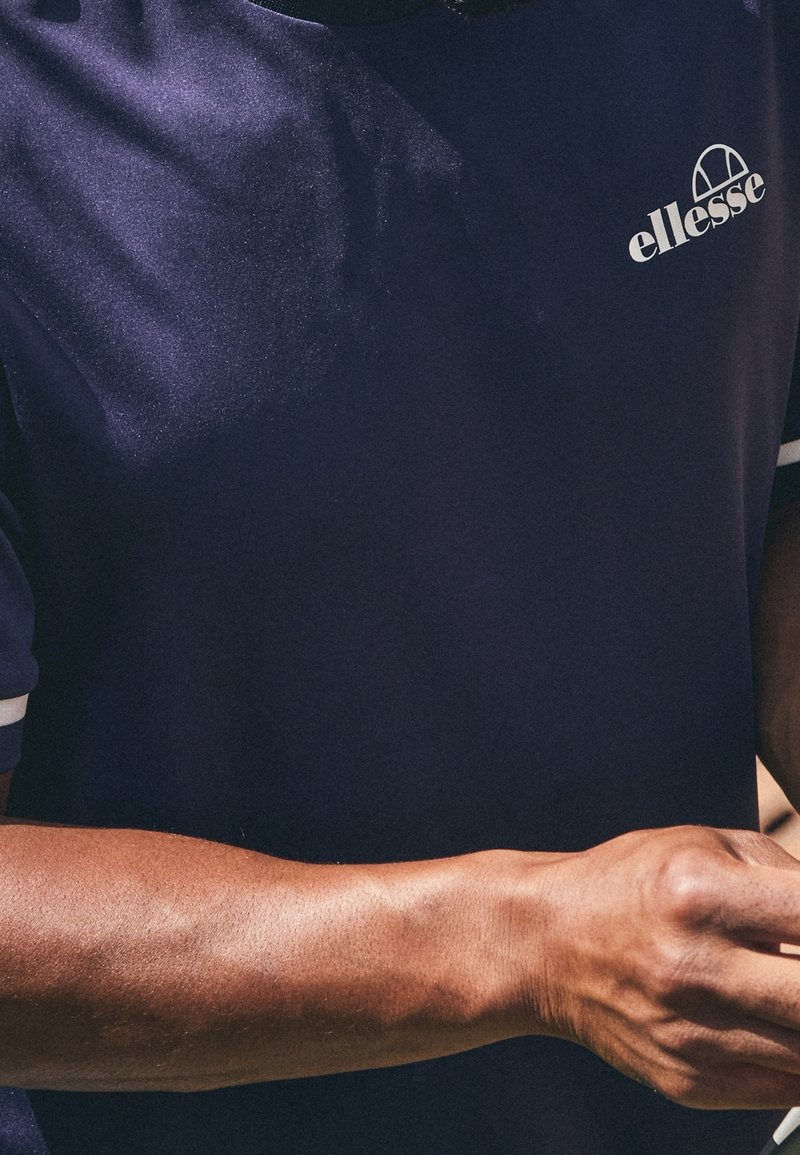 Námořnická modrá sportovní polokošile s krátkým rukávem, se bílými okraji a embosovaným logem na levé hrudi. Hladká textura, přiléhavý střih.