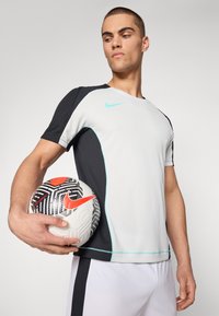 Nike urheilupaita valkoisena ja mustana, turkoosilla korostuksella, lyhyet hihat, kapea malli. Pitää kädessään kuvioitua musta-valkoista jalkapalloa.