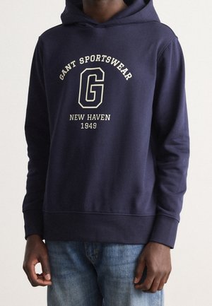 Sweat à capuche bleu marine avec le texte « GANT Sportswear », un grand « G » et « New Haven 1949 », porté avec un jean bleu clair sur une personne.
