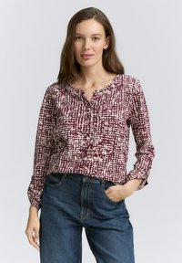 Blouse à manches longues de couleur bordeaux avec un motif texturé de points blancs et de formes irrégulières. Présente un col rond et des détails de boutons. Porté avec un jean bleu.