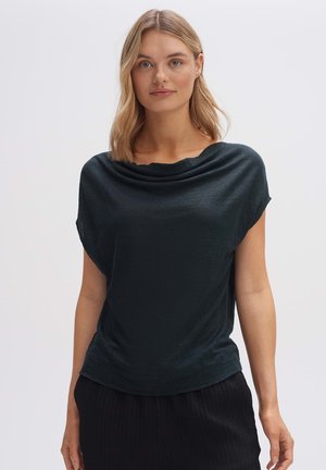 Cowl neck dark green top with soft, textured fabric; relaxed fit and short sleeves. Paired with black woven pants.