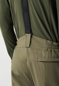 Nahaufnahme einer Person in olivgrünen Hosen mit schwarzen Hosenträgern und einem passenden olivgrünen Langarmoberteil.