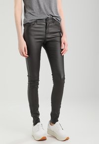 Pantalones skinny de piel sintética negra con una textura brillante, cintura alta y bolsillos delanteros funcionales, combinados con una camiseta gris y zapatillas blancas.