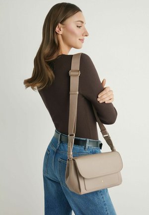 Kobieta ubrana w ciemnobrązową prążkowaną bluzkę i niebieskie dżinsy, niosąca beżową torebkę na ramię z regulowanym paskiem na ramieniu.