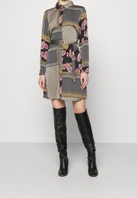 Sukienka z długim rękawem z motywem patchworkowym, zawierająca różne wzory w kolorach czarnym, złotym, różowym i białym, uzupełniona paskiem w talii.