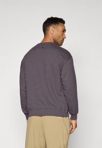 Felpa viola con scollo a girocollo, polsini e orlo a costine. Presenta un piccolo dettaglio del logo sul retro vicino allo scollo.