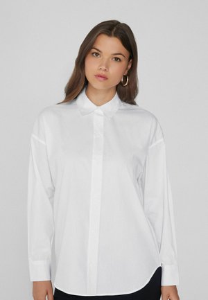Giovane donna con lunghi capelli castani che indossa orecchini a cerchio d'oro e una camicia bianca sbottonata di taglio ampio su uno sfondo grigio uniforme.