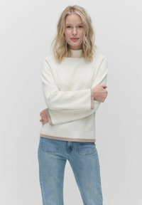 Weißer Pullover mit hohem Kragen, kontrastierenden beigen Akzenten an den Ärmeln und am Saum, aus weichem Stoff. Getragen mit hellblauen Jeans.
