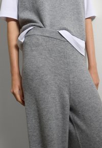 Persona che indossa un top senza maniche grigio lavorato a maglia e pantaloni abbinati sopra una maglietta bianca a maniche corte, in piedi contro un muro semplice.