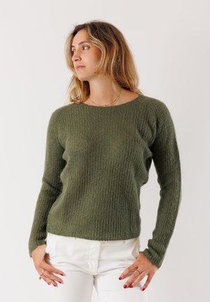 Donna con capelli biondi mossi, che indossa un maglione di lana verde oliva e pantaloni bianchi, che guarda a sinistra con le mani appoggiate sui fianchi.
