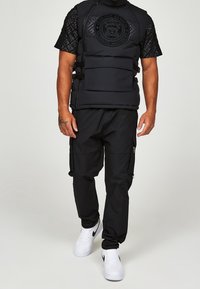 Czarna pikowana kamizelka z teksturowanym logo i przednimi kieszeniami, zestawiona z czarnymi spodniami typu cargo. Stylizację dopełniają białe trampki.