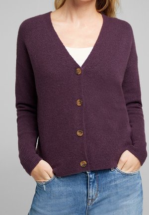 Femme portant un cardigan violet à col en V boutonné sur un haut blanc, les mains dans les poches d'un jean en denim bleu.