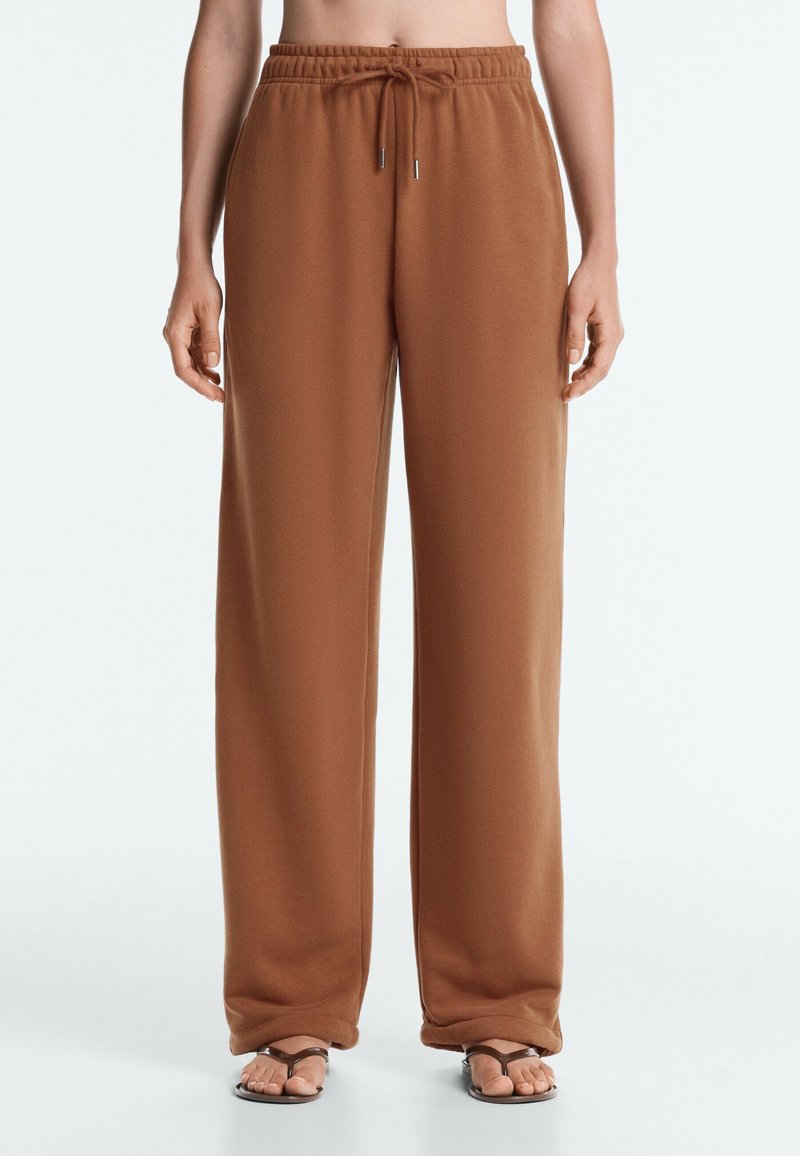 Pantalon de survêtement marron à jambes larges avec une taille élastique et un cordon, porté avec des sandales marron à brides sur une personne debout.