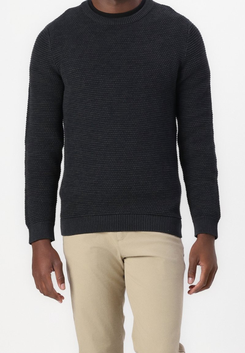 Ciemnoszary sweter z dzianiny o fakturowanym wzorze, z okrągłym dekoltem oraz ściągaczami na mankietach i dole. Połączony z jasnobeżowymi spodniami.