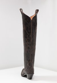 Bota de vaquero de cuero negro con punta afilada, caña alta y detalles de costura en zigzag a lo largo de los laterales. Parte superior texturizada y tacón angulado.