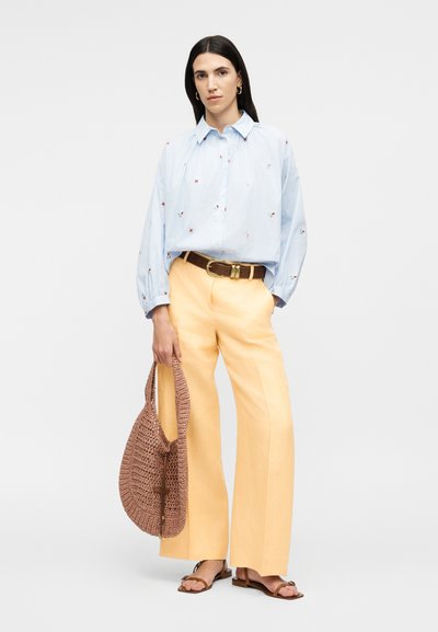 Femme aux longs cheveux bruns portant un chemisier bleu clair à petites fleurs rouges, un pantalon large jaune, une ceinture marron, des sandales, et tenant un sac tressé marron.