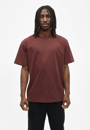 Man met dreadlocks die een eenvoudige donkerbruine korte mouw t-shirt en zwarte broek draagt, staand tegen een witte achtergrond.