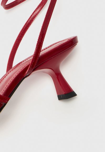 Czerwone lakierowane sandały na wysokim obcasie z cienkimi paskami, konturową sylwetką oraz trójkątnym obcasem na czarnej podstawie.