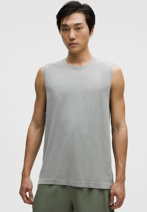 Junger Mann mit glatten schwarzen Haaren, der ein hellgraues ärmelloses Shirt und olivgrüne Shorts vor einem schlichten Hintergrund trägt.