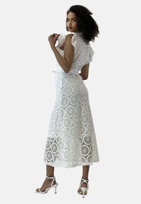 Abito midi bianco con ricami in pizzo floreale, dettagli arricciati sulle spalle, una cintura da annodare e una silhouette fluida. Indossato con tacchi aperti.