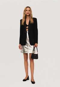 Blazer nero sopra un bralette nero, mini gonna in metallo argentato, borsa nera con hardware dorato e scarpe flat nere. Texture lisce in tutto.