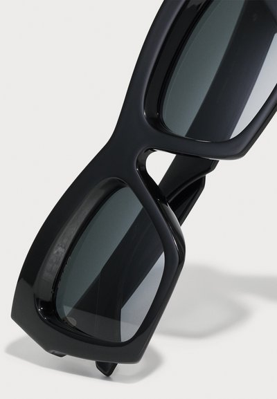 Črne sončne očala z masivnim okvirjem, kvadratnimi lečami in gladkim, sijočim zaključkom. Imajo edinstveno obliko izreza na okviru.