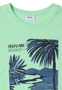 Chemise vert menthe avec une illustration noir et bleu de palmier et d'océan, comportant un texte sur le soin de soi et le bien-être en tant qu'investissement et style de vie.