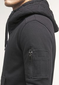 Felpa nera con cappuccio trapuntato, caratterizzata da una tasca con zip sulla manica sinistra e tessuto texturizzato, che mette in evidenza un design aderente.
