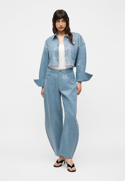 Γυναίκα που φοράει ανοιχτό μπλε τζάκετ με παγιέτες, cropped, και αντίστοιχο παντελόνι με φαρδύ πόδι, με μαύρα σανδάλια με τακούνι, στέκεται μπροστά σε λευκό φόντο.