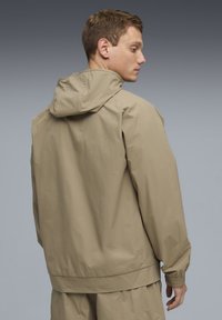 Μπεζ μπουφάν με κουκούλα, κατασκευασμένο από λείο, ελαφρύ υλικό. Διαθέτει ελαστικά μανίκια και τελείωμα, με μινιμαλιστικό σχέδιο και χωρίς ορατή σήμανση.