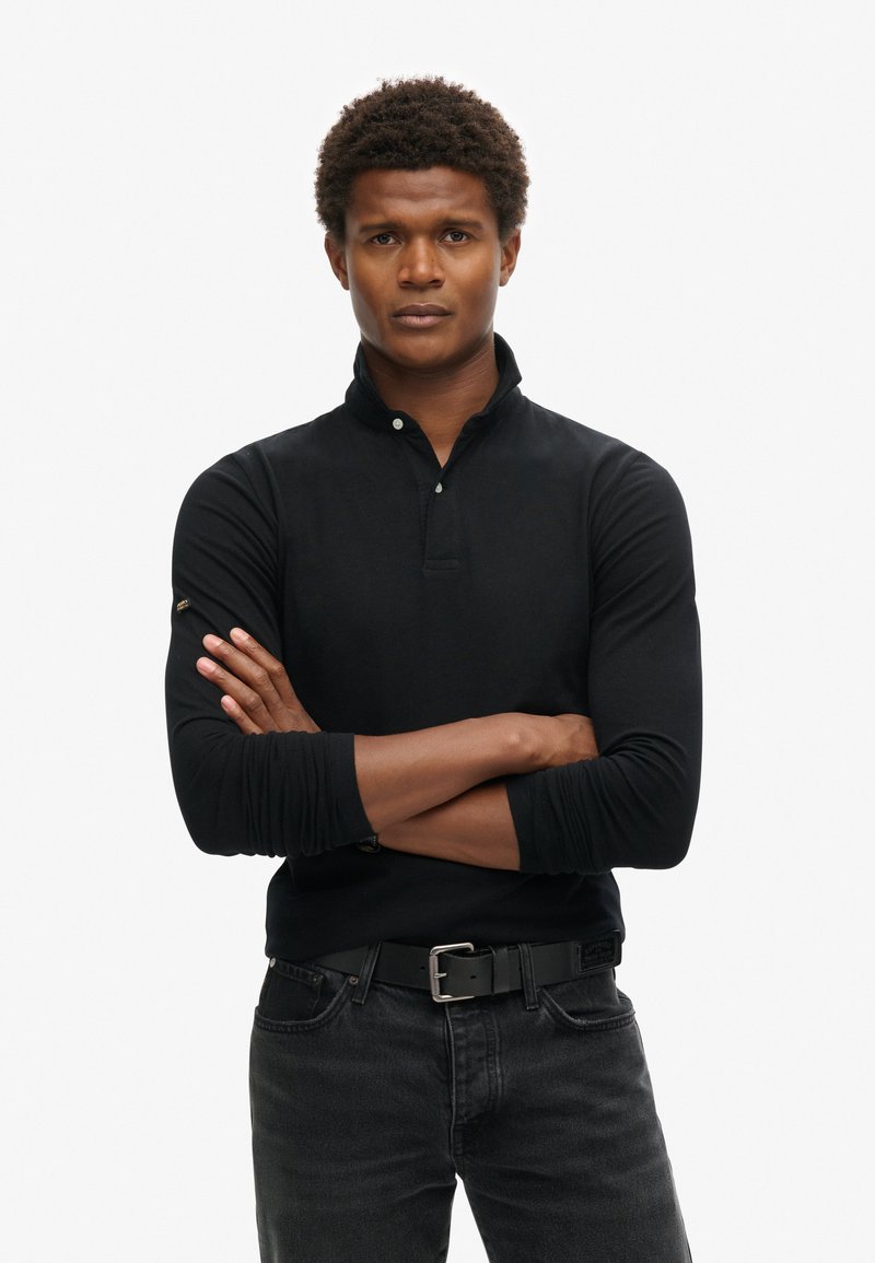 Camisa polo negra de manga larga con cuello, que presenta un cuello abotonado y puños. Combinada con jeans de mezclilla oscuros y un cinturón negro.