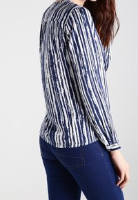 Maglietta a righe blu e bianca a maniche lunghe con vestibilità morbida, caratterizzata da un colletto rotondo e una texture di tessuto morbida. Indossata con jeans blu.