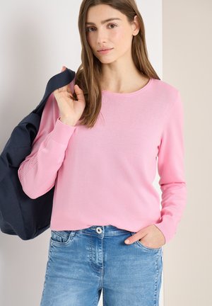 Młoda kobieta z długimi brązowymi włosami, ubrana w różowy sweter i niebieskie dżinsy, trzymająca ciemną kurtkę przewieszoną przez ramię.