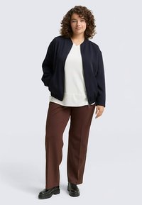 Giubbotto bomber blu navy con scollo rotondo, abbinato a una maglietta bianca e pantaloni marroni. Tessuto liscio con vestibilità comoda e polsini elastici.