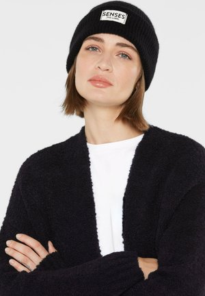 Black ribbed knit beanie with a label; soft-textured black cardigan layered over a white shirt. Neutral tones, relaxed fit.