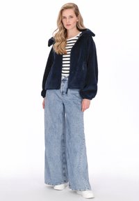 Granatowa kurtka polarowa z kapturem, z zamkiem błyskawicznym z przodu i kieszeniami, połączona z jasnoniebieskimi szerokimi spodniami z wysokim stanem i białymi butami.