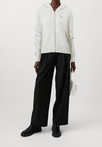 White zip-up hoodie with a kangaroo pocket and hood, paired with black pinstriped wide-leg pants and black ankle boots, holding a white bag.