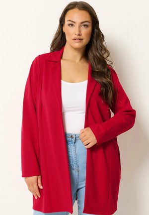 Vrouw met lang bruin haar die een rode open blazer draagt over een wit topje en lichtblauwe jeans, staand tegen een effen achtergrond.
