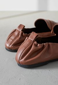 Ballerine in pelle marrone con dettaglio arricciato sulle punte, dotate di una texture morbida e suola in gomma nera bassa. Forma arrotondata con un design minimale.