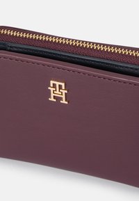 Burgunderfarget lærpung med logo i gullhardware og glidelås på toppen. Glatt tekstur med kontrasterende svart innside.