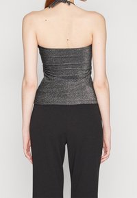 Kobieta w błyszczącej topless z wiązanym halterem, w zestawieniu z czarnymi spodniami o wysokim stanie, pokazana z tyłu.