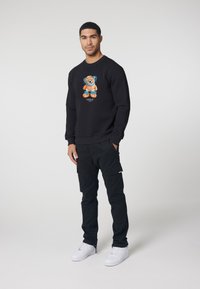 Black sweatshirt with a colorful teddy bear graphic on the front, paired with black cargo pants and white sneakers. Simple, casual design.