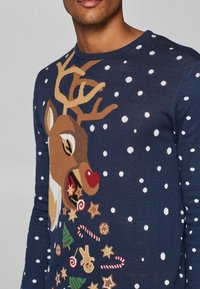 Marineblauer Strickpullover mit einem Rentier-Motiv, das Geweihe und eine rote Nase zeigt, umgeben von weißen Schneeflocken und festlichen Dekorationen.