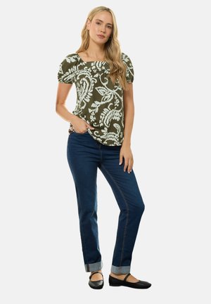 Kvinde iført en olivengrøn mønstret bluse, mørke opbukte jeans og sorte flade sko, stående med den ene hånd i lommen mod en hvid baggrund.