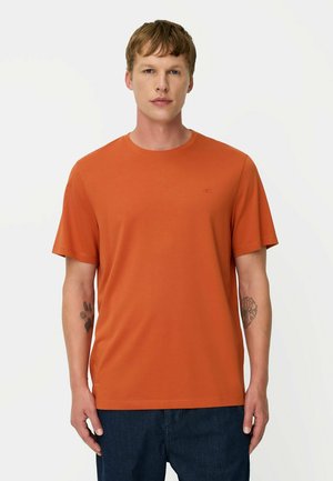 Mann mit kurzen hellbraunen Haaren, der ein oranges kurzärmeliges T-Shirt und dunkelblaue Hosen trägt, steht vor einem schlichten weißen Hintergrund.