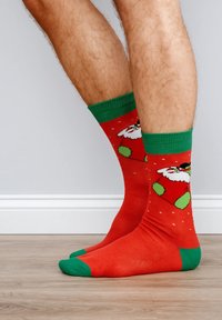 Rote Socken mit grünen Akzenten und einem Weihnachtsmann-Motiv. Das Material wirkt weich mit glatter Textur. Kontrastfarben an Zehen und Bündchen.