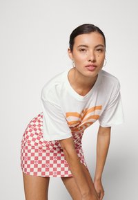 Camiseta branca com design abstrato laranja; saia mini xadrez rosa com padrões florais; modelo posa com as mãos nas coxas.