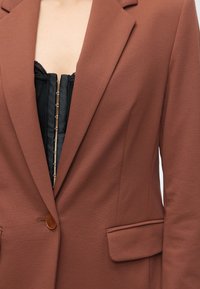 Femme portant un blazer marron à un bouton sur un corset noir sans bretelles avec des crochets à l'avant, montrant partiellement la poitrine et le cou.