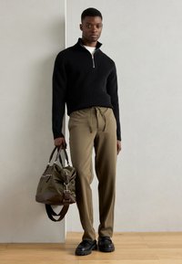 Pull en maille côtelée noir avec zip, pantalon beige à cordon de serrage, chaussures noires épaisses, portant un sac vert olive avec des accents marron et des fermetures éclair.